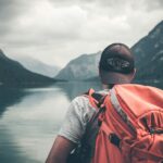 man with red hiking backpack facing body of water and mountains at daytime