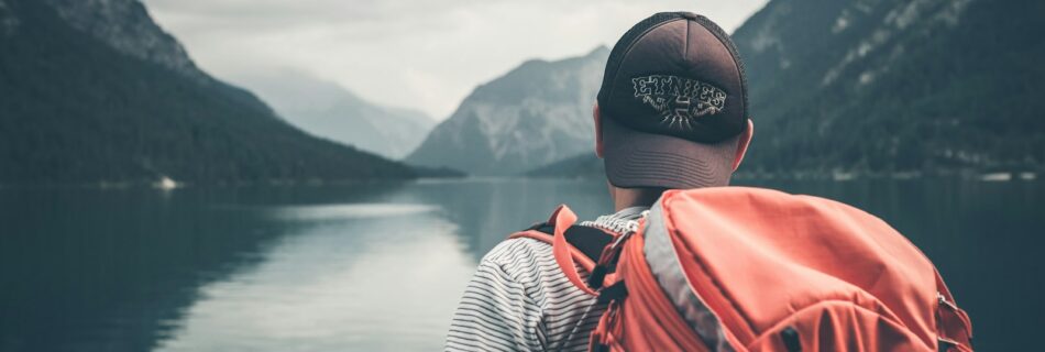 man with red hiking backpack facing body of water and mountains at daytime
